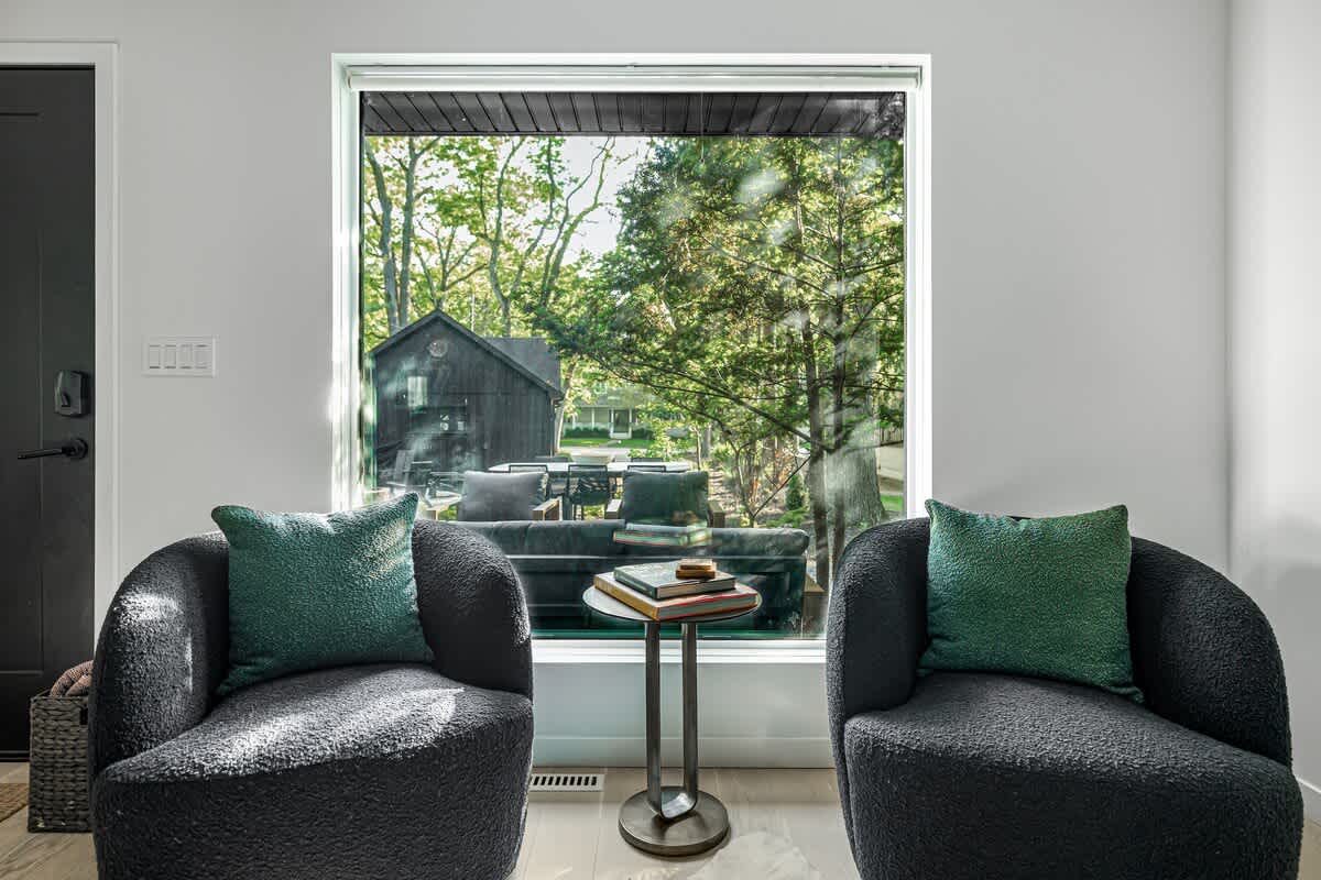 Two dark gray boucle armchairs with green throw pillows flank a small round side table with books, facing a large window that offers a view of lush green trees and a dark shed outside.