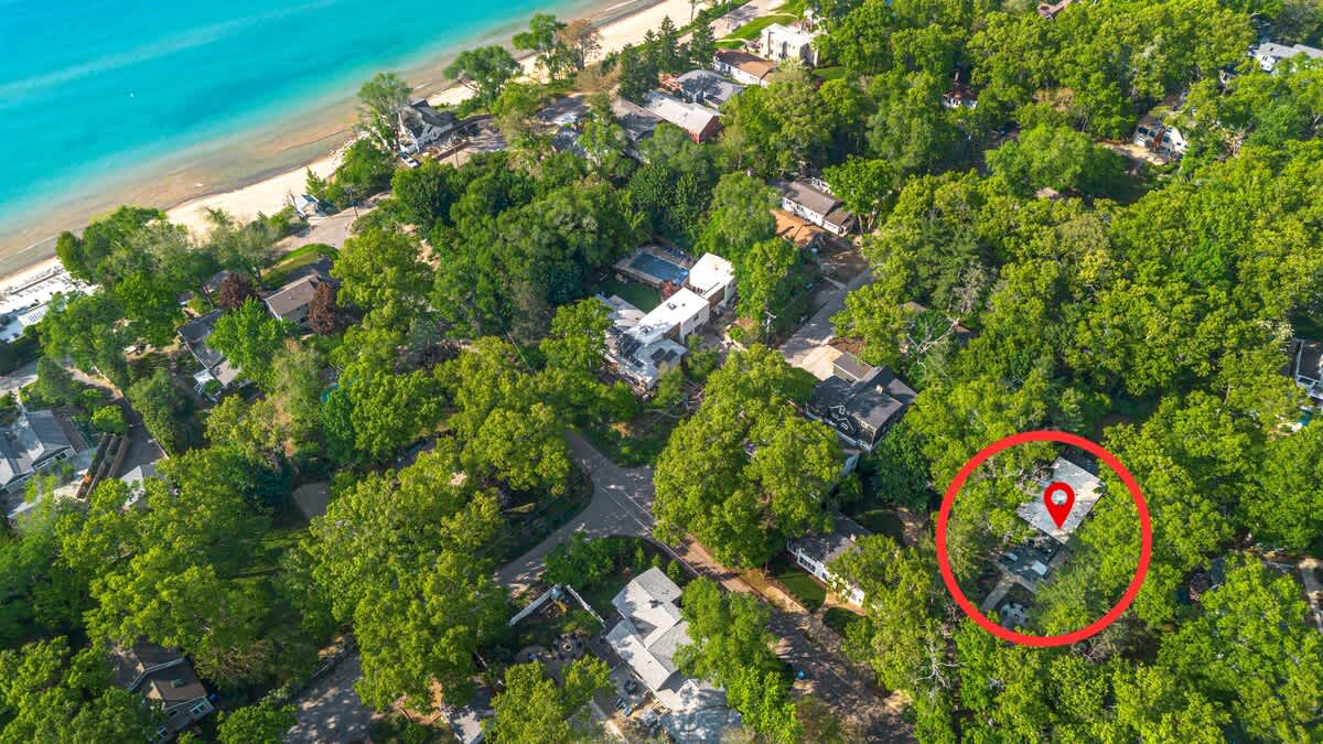 A wide aerial shot highlights the property, marked by a red pin, nestled within a dense green forest, with a beautiful blue lake or ocean and sandy beach visible in the upper left background.