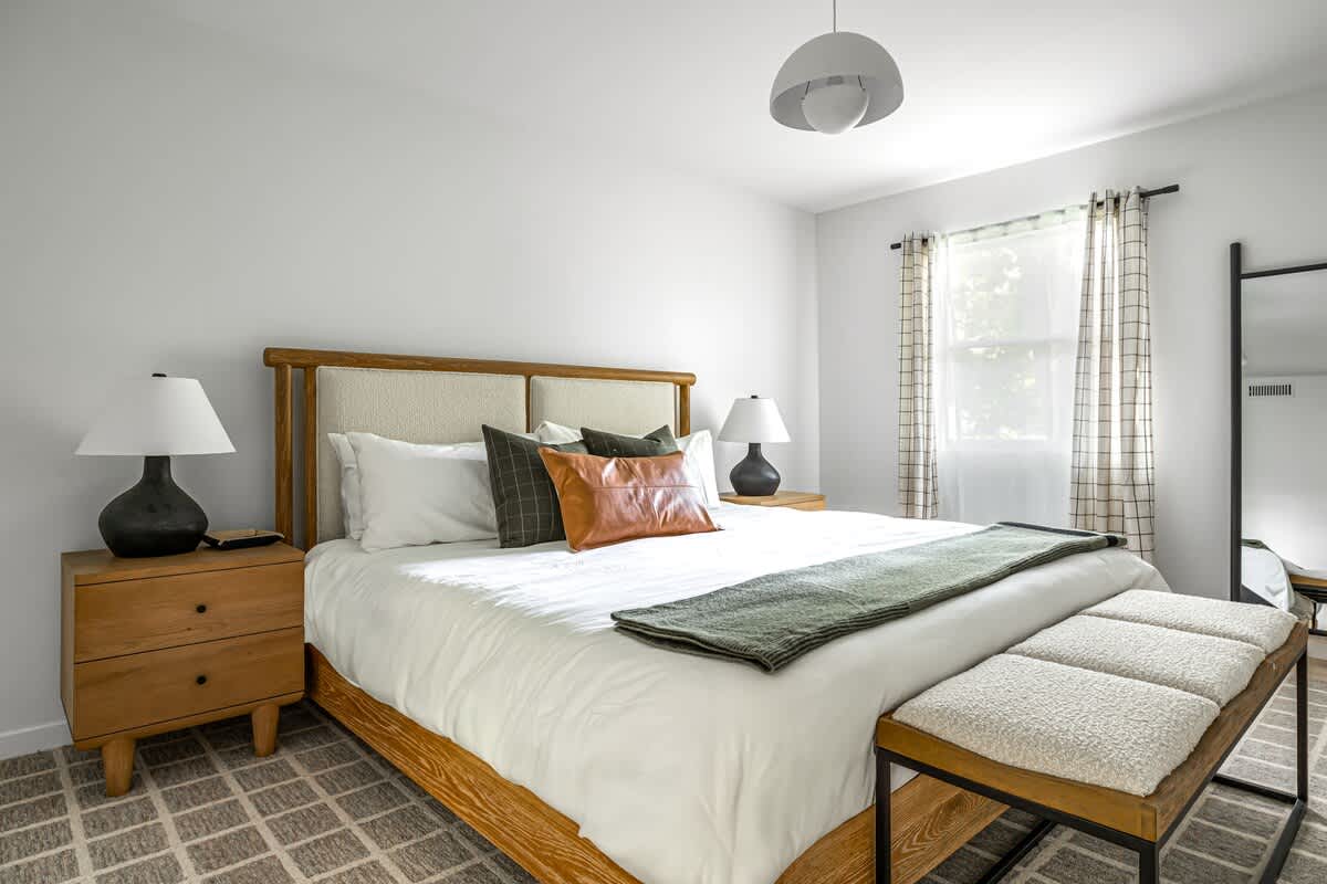 Cozy bedroom featuring a queen-sized bed with a wooden and upholstered headboard, white bedding, and green and brown accent pillows. Two wooden nightstands with black lamps flank the bed, with a bench at the foot.