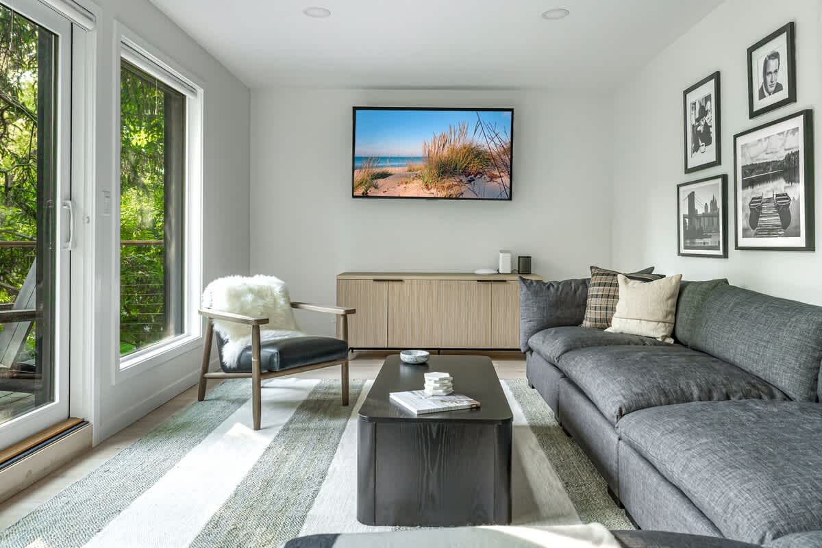 Cozy living room featuring a wall-mounted TV above a light wood media console, a dark gray sectional sofa, and a black coffee table. A black leather armchair with a white faux fur throw sits by a large glass door.
