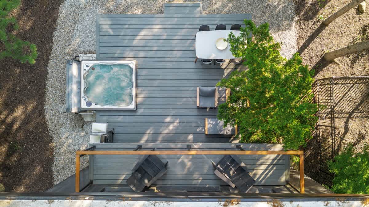 An aerial view shows a large grey composite deck featuring a bubbling hot tub, an outdoor dining table with chairs, and additional lounge seating, surrounded by gravel and lush green trees.
