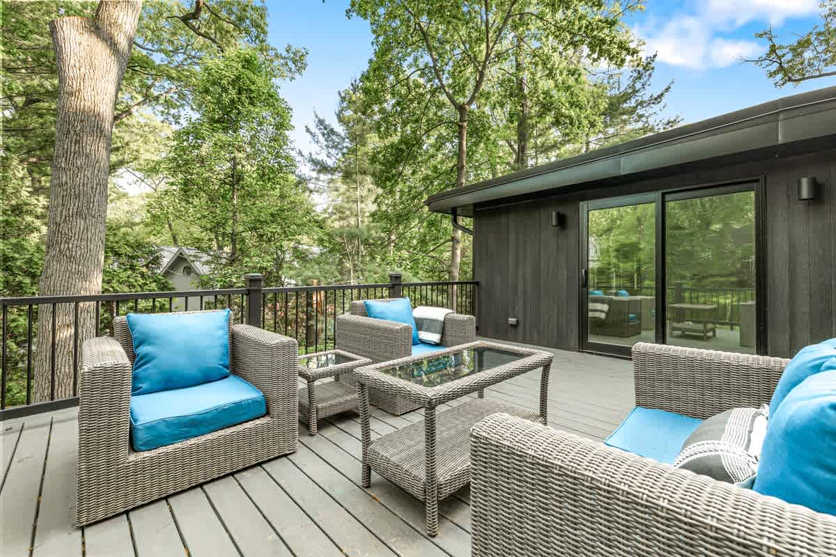 Elevated balcony with grey wicker outdoor seating and bright blue cushions, offering a view of surrounding green trees. The modern black exterior of the house is visible through sliding glass doors.