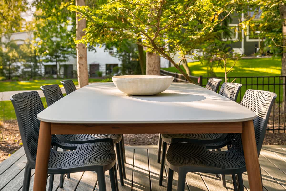 Outdoor dining area on a wooden deck, featuring a long light-colored table with dark modern chairs, surrounded by green trees and bathed in natural sunlight.