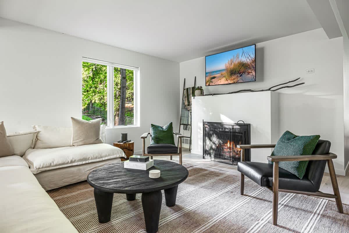 Bright living room with a large white sectional sofa, a dark round coffee table, and a white fireplace with a wall-mounted TV above. A large window provides views of the surrounding trees.