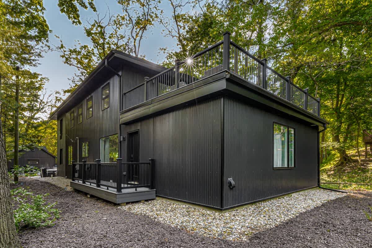 The modern black-sided house features corrugated metal siding and large windows, with a ground-level deck and an upper-level deck with railings, surrounded by gravel and natural landscaping under a bright sky.