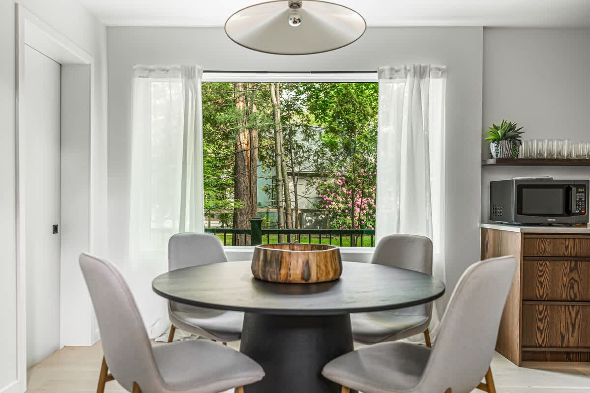Inviting breakfast nook with a round black table, four light grey chairs, and a large window offering lush green tree views, illuminated by a modern pendant light.