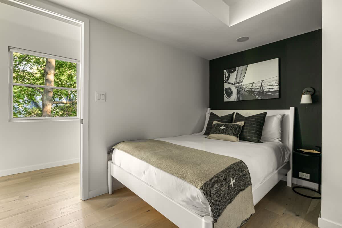 View of one bed in a shared bedroom, featuring a white bed frame, white bedding, and plaid accent pillows. A black accent wall behind the bed showcases a large black and white sailboat artwork.