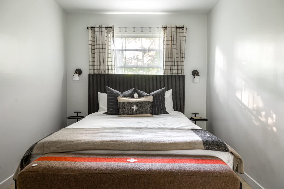 View of the second bed in a shared bedroom, featuring a dark upholstered headboard, white bedding, and plaid accent pillows. Two wall sconces flank the bed, with a window above.