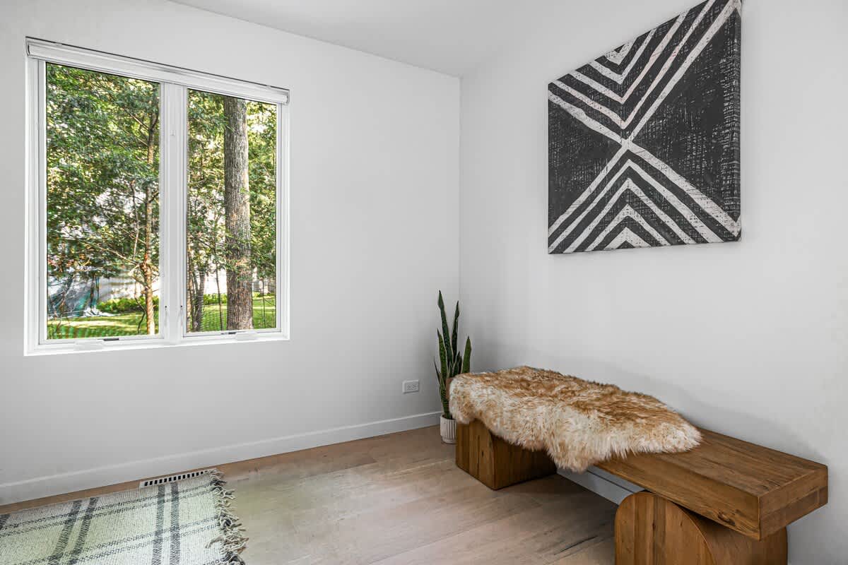 A minimalist room with light wood floors, a large window overlooking green trees, and a wooden bench adorned with a faux fur throw. A tall snake plant sits beside the bench, and a geometric black and white artwork hangs on the wall.