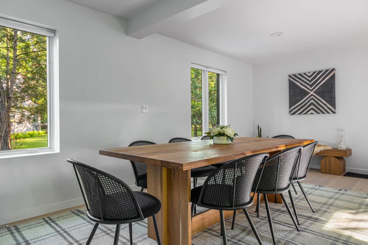 Modern dining area with a large natural wood table, black woven chairs, and a plaid area rug, set against white walls with two large windows and abstract wall art.