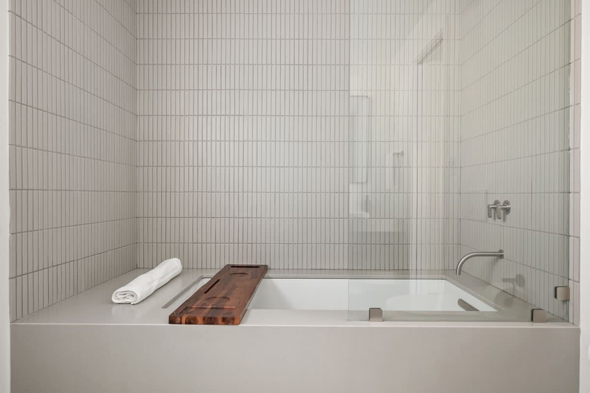 Close-up of a modern deep soaking tub with a wooden bath caddy and a white towel, set against light grey vertical tile walls and a clear glass shower screen.
