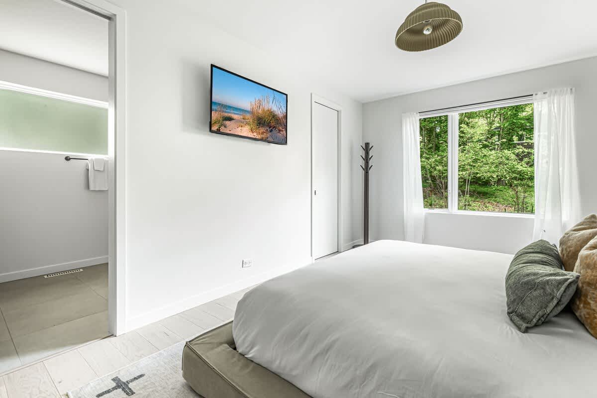 Bright bedroom with a large bed featuring white bedding and decorative pillows, a wall-mounted TV, and a window overlooking lush green trees, creating a serene and modern space.