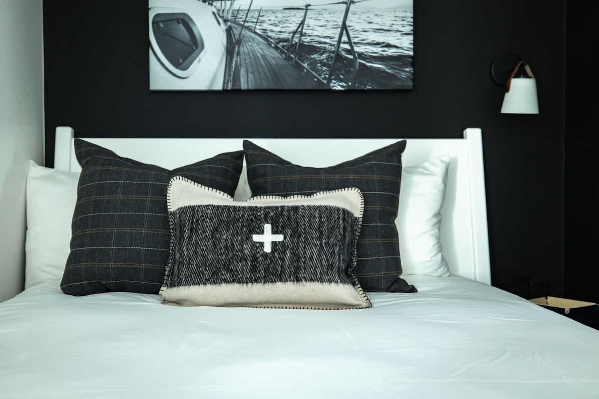 Close-up of a bed with white bedding and a collection of plaid and textured accent pillows, including one with a white cross design. A black accent wall is visible behind the bed.