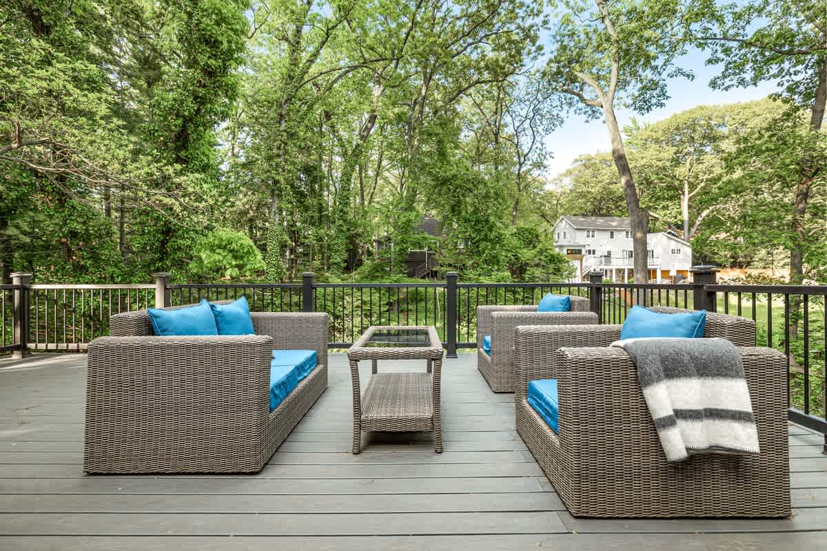 A spacious outdoor deck features modern wicker-style lounge chairs with bright blue cushions, arranged around a matching coffee table, all set against a lush green forest backdrop with a distant house visible.