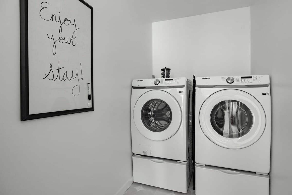 Dedicated laundry room with a white front-loading washer and dryer stacked on pedestals, set against a white wall with a framed 'Enjoy your Stay!' sign.