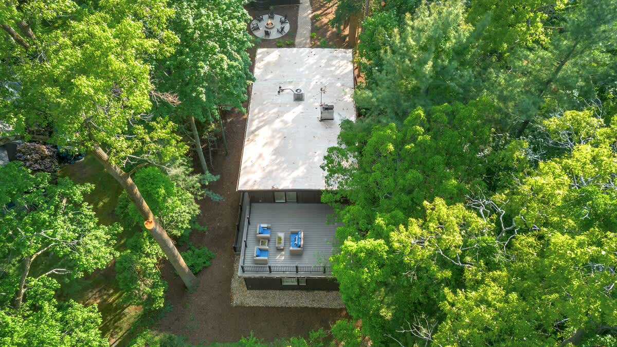 A high aerial view captures the long, rectangular house with a light-colored roof, nestled among dense green trees, showcasing the outdoor deck with seating and a hot tub, and a fire pit area further down a path.