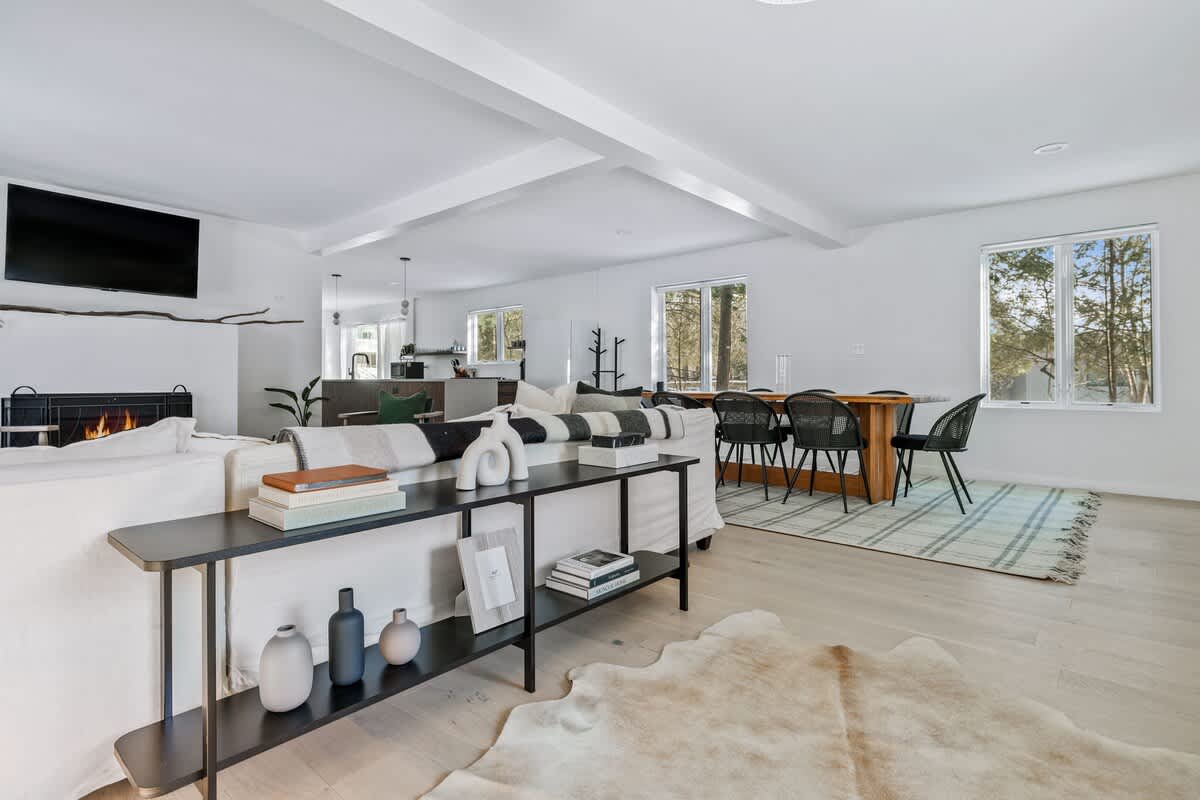 Bright open-concept living and dining area featuring light wood floors, white walls, a modern fireplace with a TV above, and a long black console table behind a white sofa.