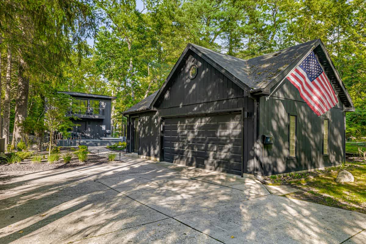 Dark-colored modern house with a detached garage, both featuring black siding and surrounded by tall green trees. An American flag hangs from the garage, and a concrete driveway leads to the structures.