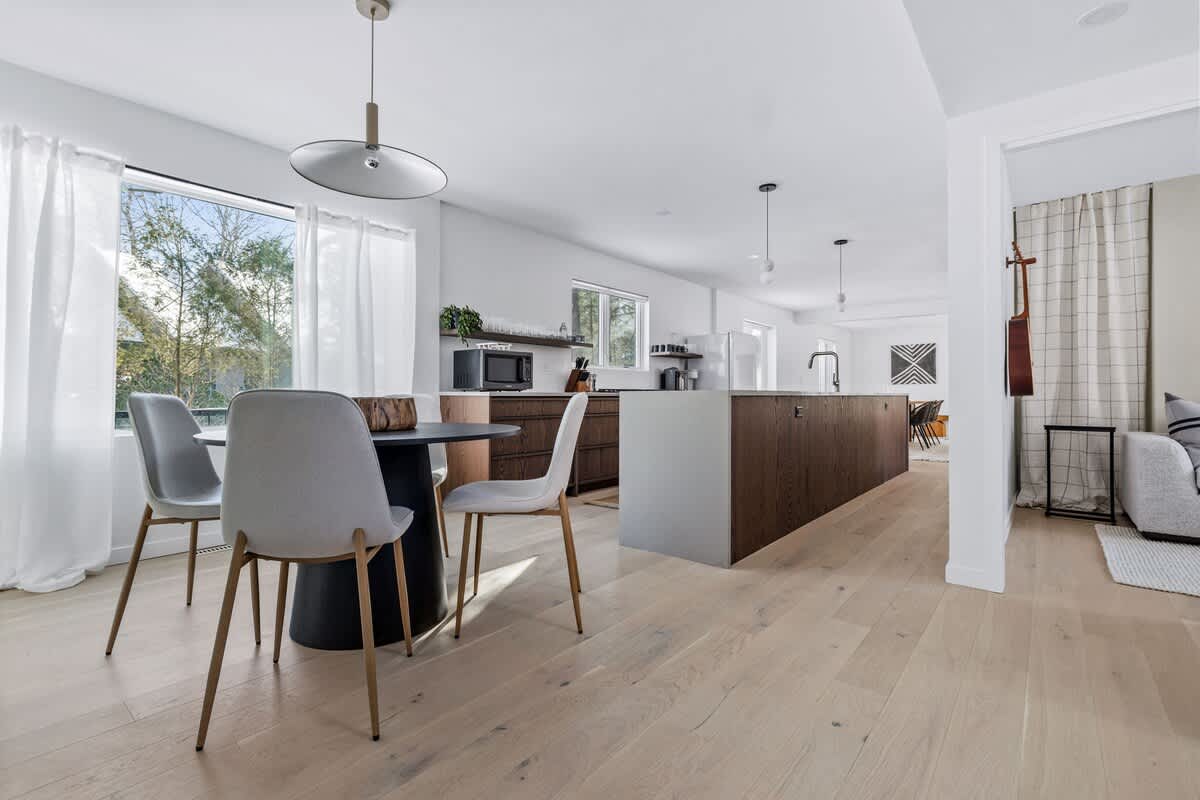 Modern kitchen with a large island and dark wood cabinetry, adjacent to a bright breakfast nook featuring a round black table and light grey chairs by a large window with white curtains.