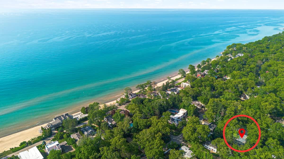 Aerial view showcasing a lush green forested area bordering a vibrant blue lake or ocean, with a sandy beach shoreline. A red pin highlights the property nestled among the trees.