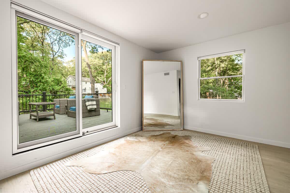 Bright, open interior space with light hardwood floors and a large patterned rug, featuring a sliding glass door that opens to a deck with outdoor seating and lush green trees. A tall mirror leans against the wall.