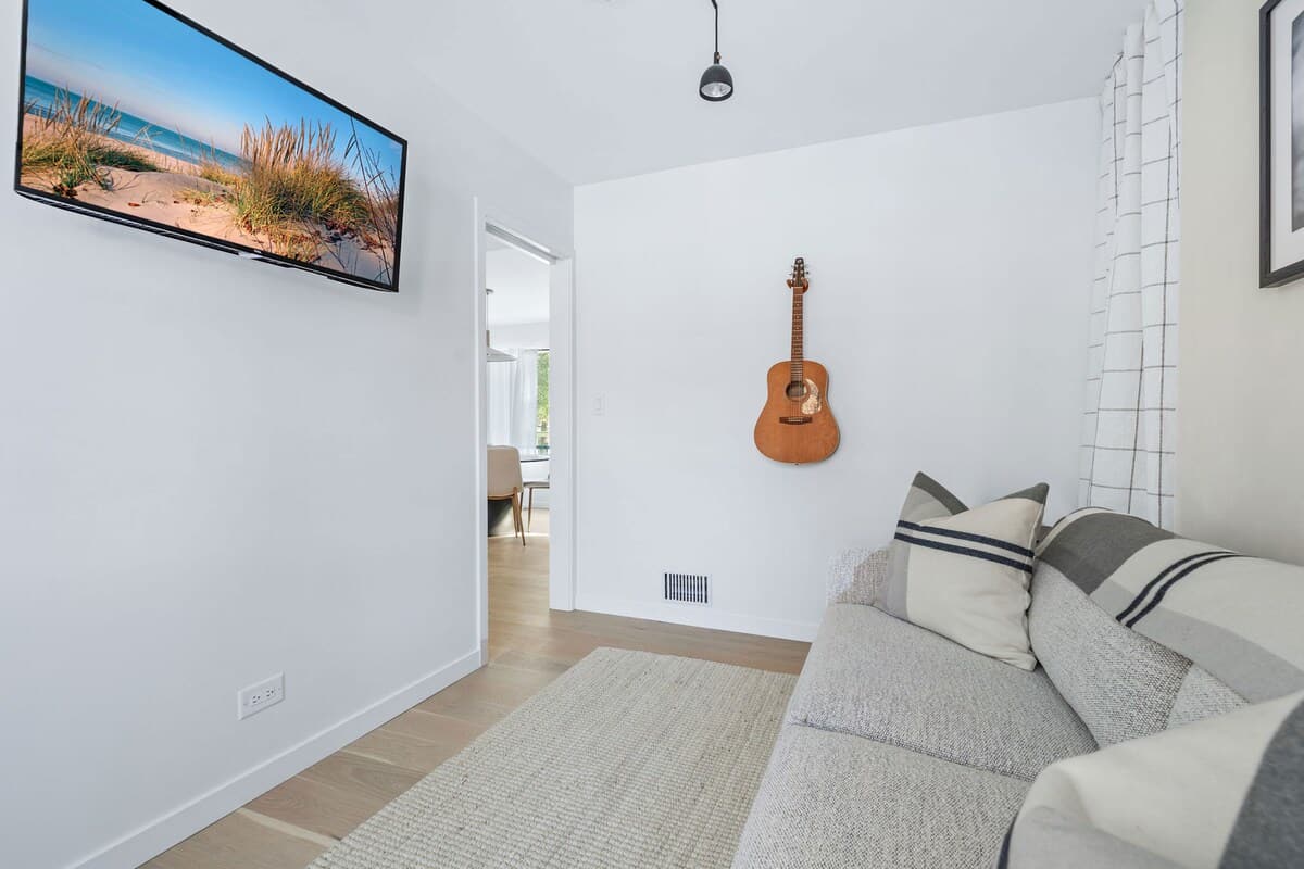 A narrow living space with light wood floors and a light gray sofa adorned with striped pillows and a plaid throw. A guitar hangs on the white wall, and a wall-mounted TV is visible in the distance.
