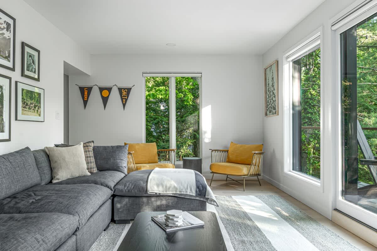 Spacious living room with a large gray sectional sofa, a striped area rug, and two yellow accent chairs by a window. Large windows and a glass door offer views of lush greenery outside.