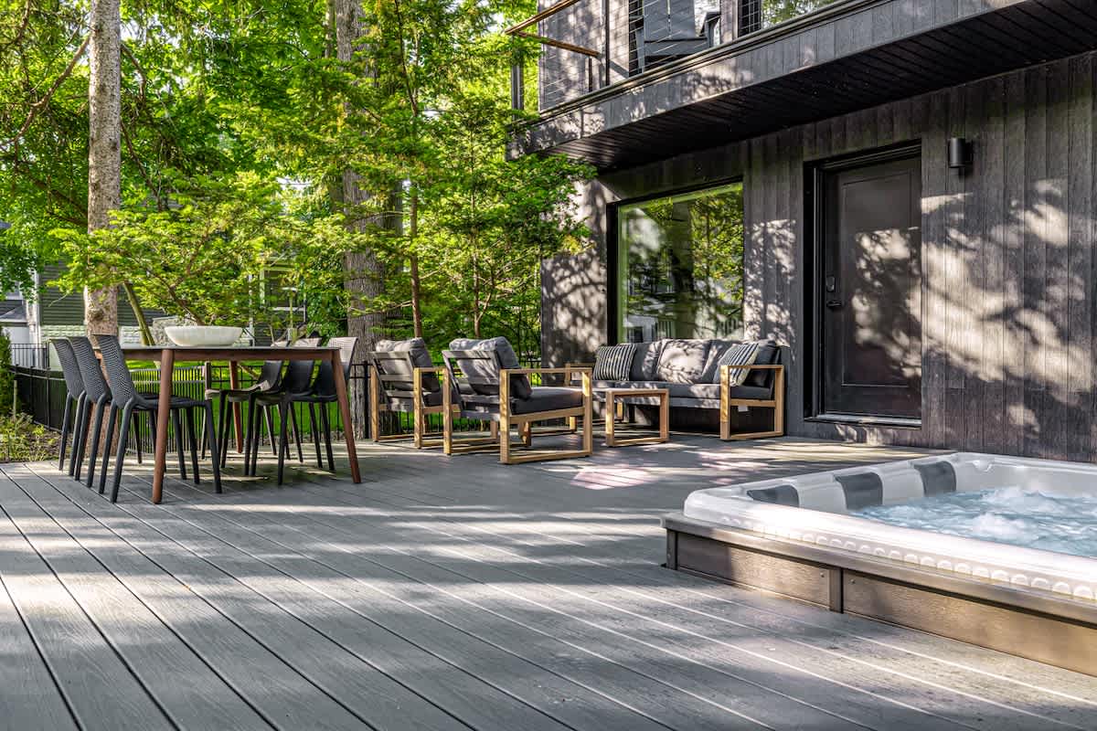 Spacious grey composite deck featuring an outdoor dining table with black chairs and a lounge area with modern outdoor sofas. A hot tub is partially visible on the right, all surrounded by lush greenery.