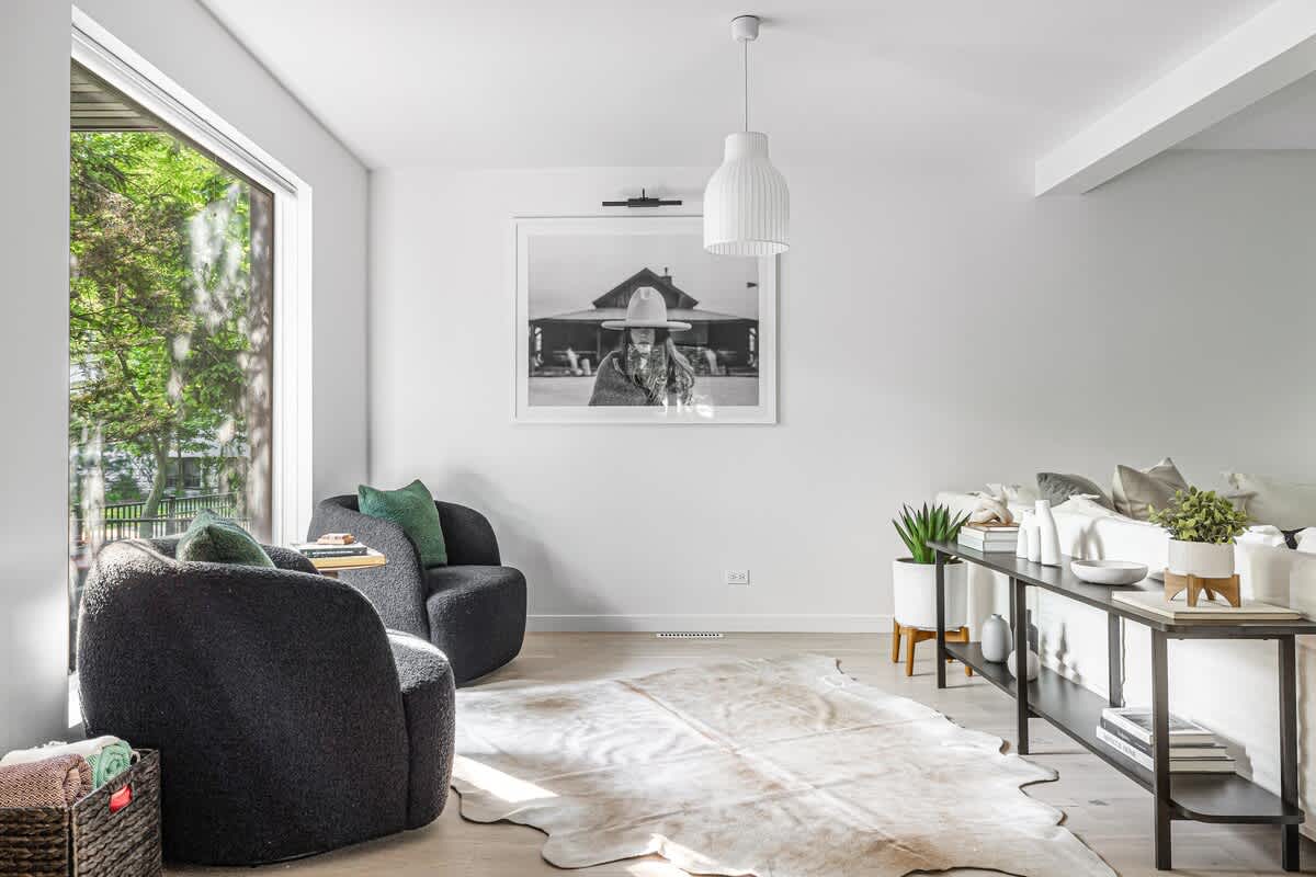 Bright living room with a large cowhide rug on light wood floors, two dark gray boucle armchairs with green pillows, and a long black console table with plants. A large black and white framed art piece hangs above the console.