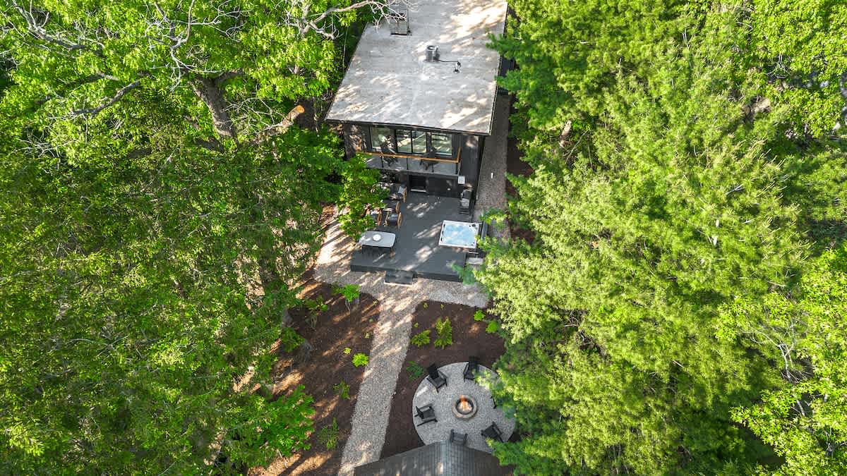 An aerial shot reveals the black-sided house with its outdoor deck, hot tub, and dining area, connected by a gravel path leading to a circular fire pit surrounded by chairs, all enveloped by lush green trees.
