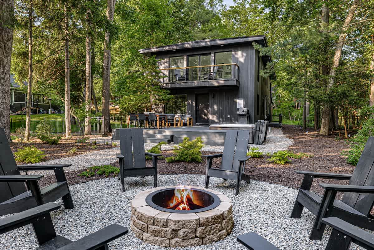 Modern black house with a second-story balcony, surrounded by lush green trees. A stone fire pit with black Adirondack chairs sits on a gravel patio in the foreground.