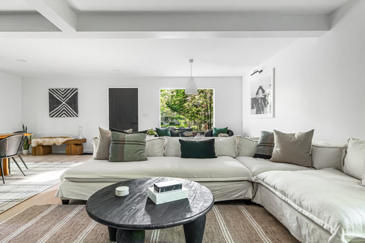 Expansive living room featuring a large white sectional sofa, a dark round coffee table, and a striped area rug. The open-concept space includes a dining area in the background and large windows offering natural light.