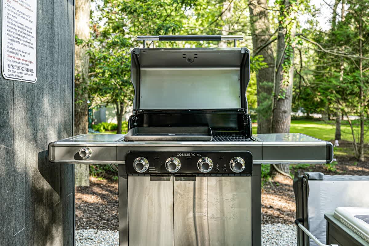Stainless steel BBQ grill with multiple burners and side tables, positioned on a gravel area next to a wooden deck, surrounded by green foliage.