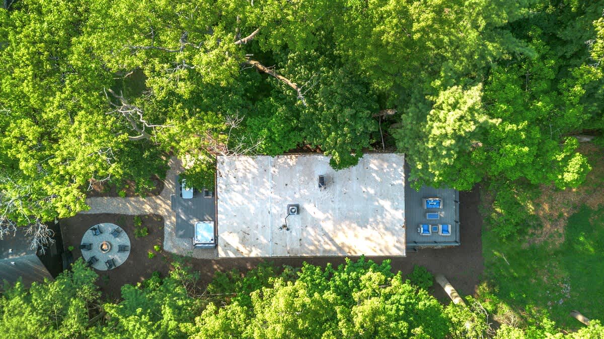 A direct overhead aerial view showcases the light-colored roof of the house, with a hot tub visible on a deck to the side, and a circular fire pit area with seating nestled among the dense green tree canopy.