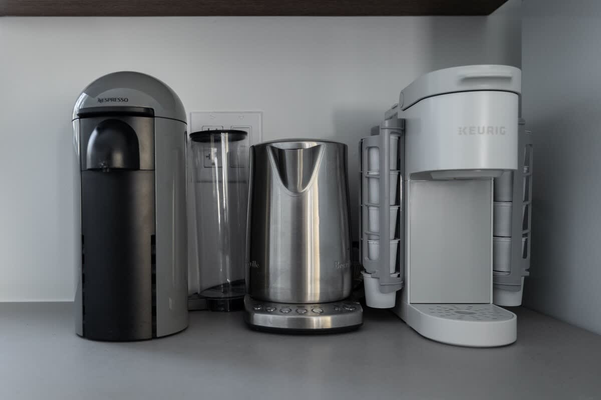 A modern coffee station featuring a Nespresso machine, a stainless steel electric kettle, and a Keurig coffee maker, all arranged on a light gray countertop against a white wall.