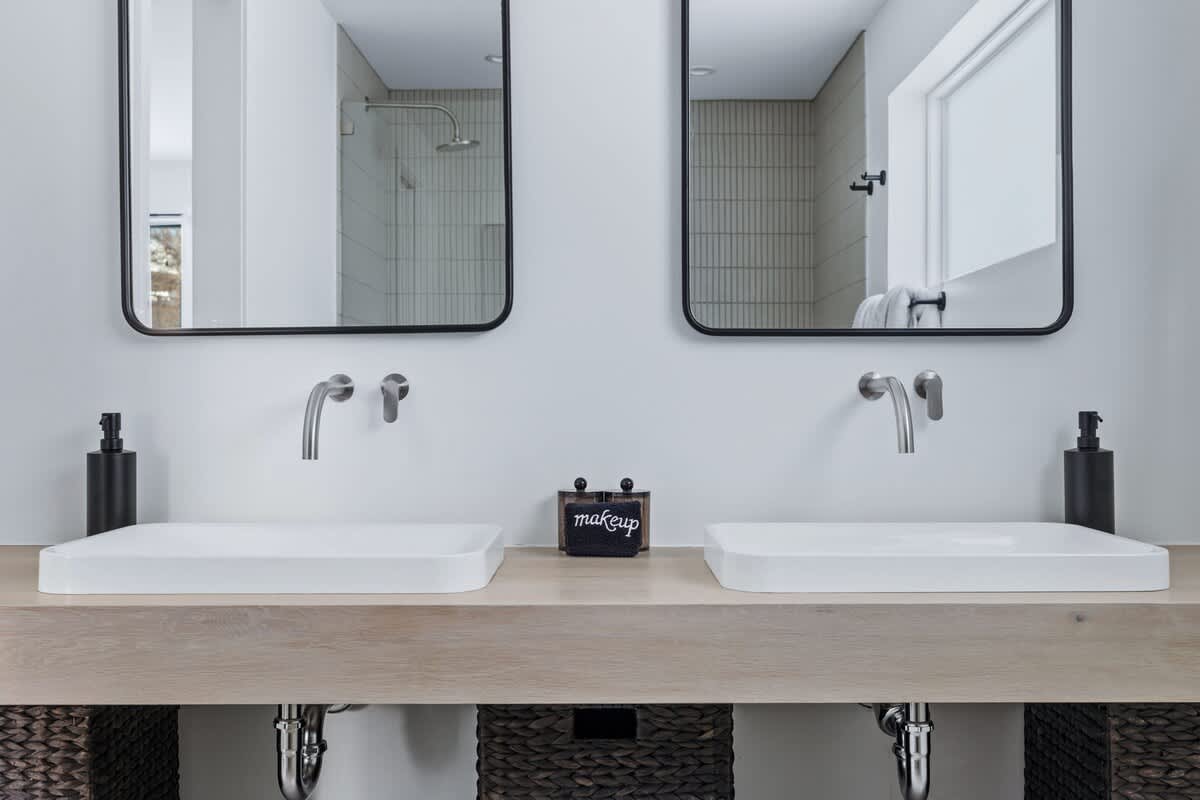 Modern double vanity area featuring two rectangular sinks, individual wall-mounted faucets, and large framed mirrors above a light wood countertop with woven storage baskets below.