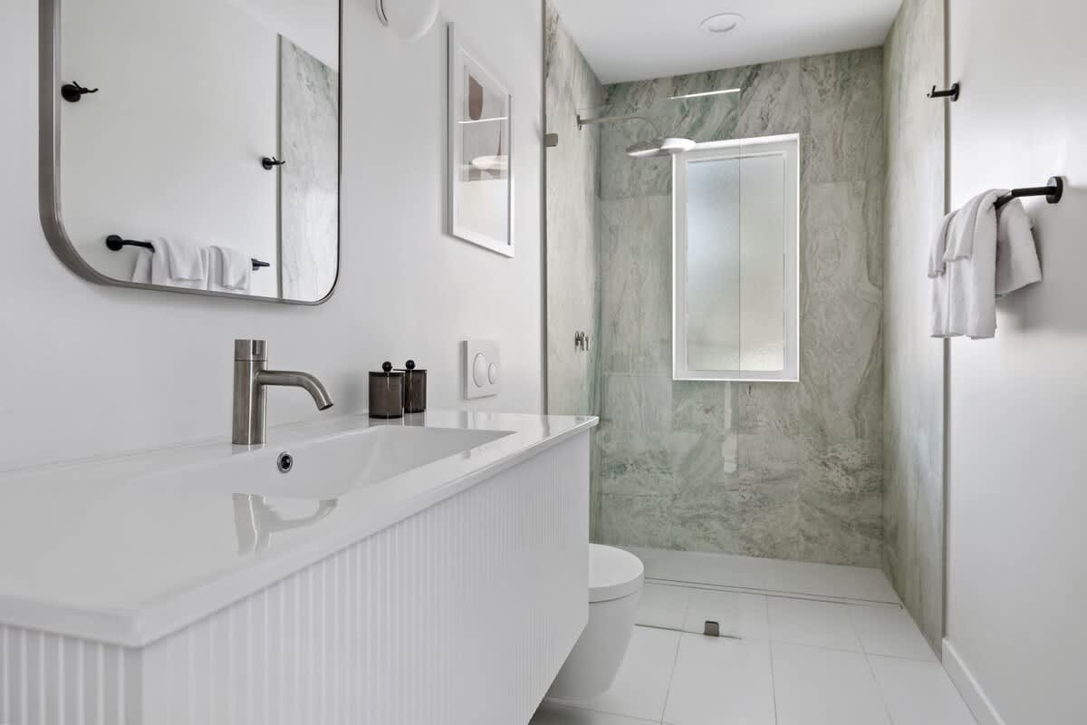 Sleek bathroom with a white fluted vanity and integrated sink, a large mirror, and a walk-in shower with green marble-patterned tile walls and a rain shower head.