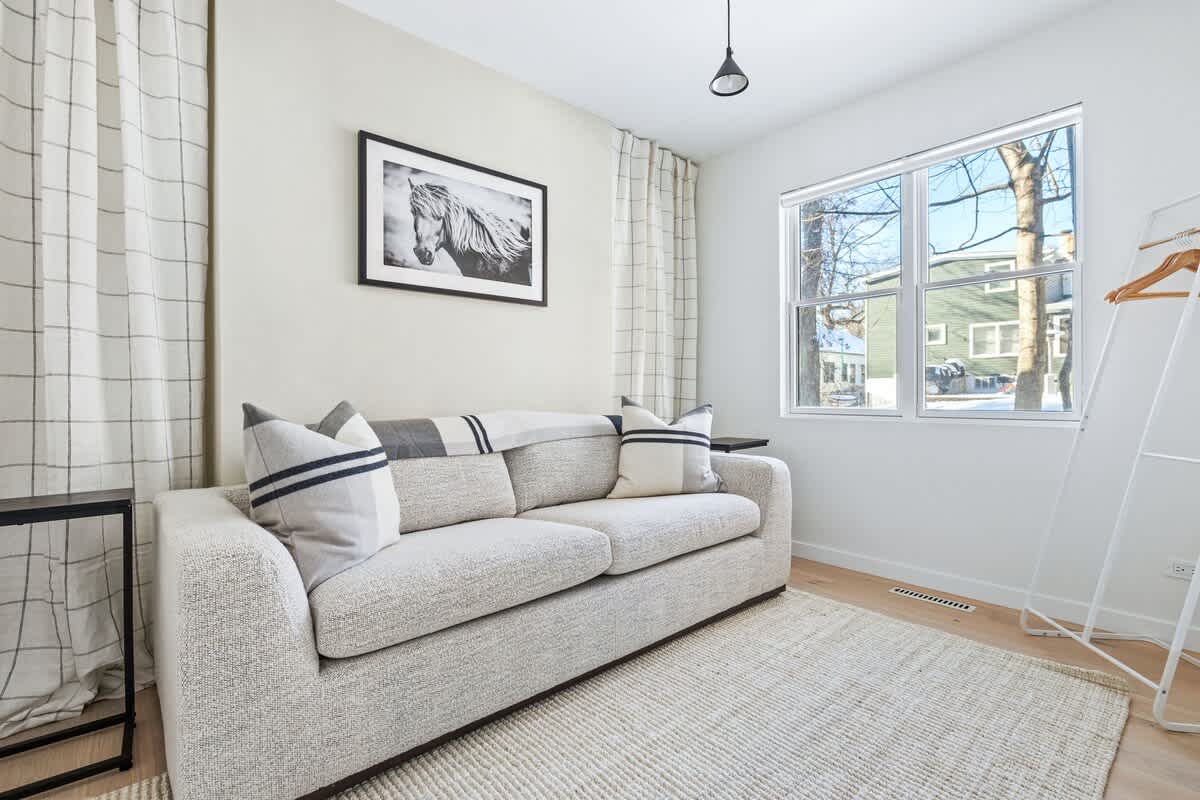 Bright bedroom with a light gray sofa, striped throw pillows, and a plaid blanket, positioned against a light-colored wall with a framed horse artwork. A large window overlooks a residential area, and a white clothing rack stands nearby.