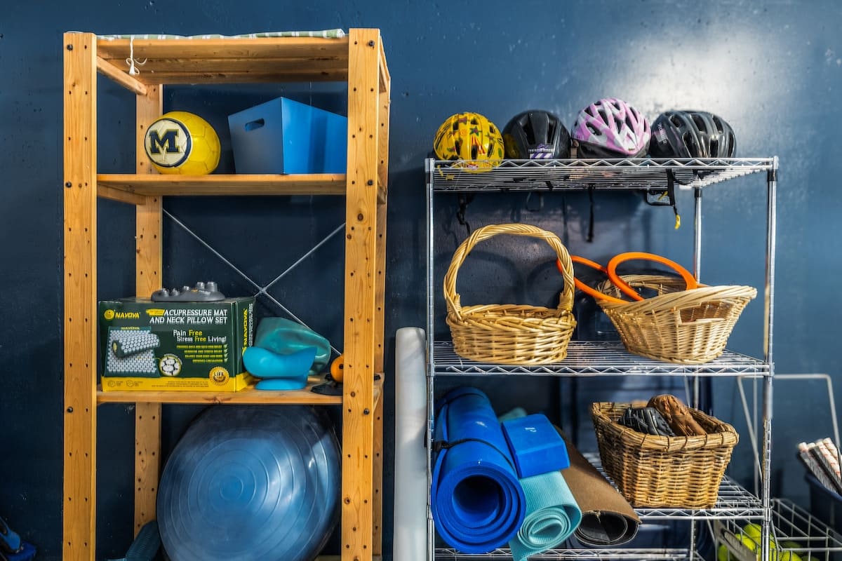Close-up of organized storage shelves in a game room, featuring a wooden unit with exercise balls and a metal shelf holding yoga mats, woven baskets, and colorful bicycle helmets.
