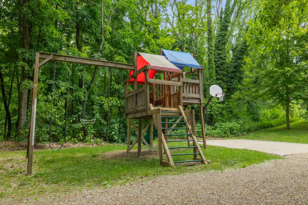 A rustic wooden playset with swings, a slide, and a small basketball hoop, situated on a grassy area next to a gravel path, surrounded by tall, leafy green trees.