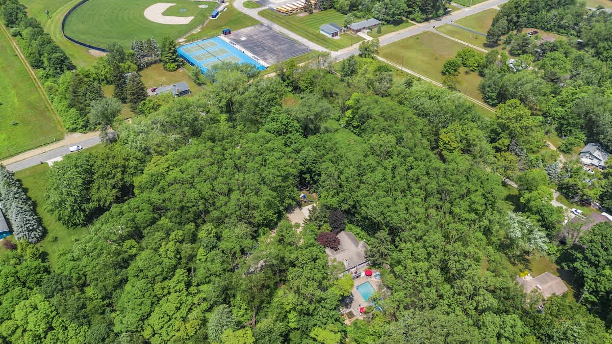 Wide aerial view showcasing a house and pool nestled within a dense forest, with a baseball field and blue tennis courts visible in the upper background, suggesting nearby recreational amenities.