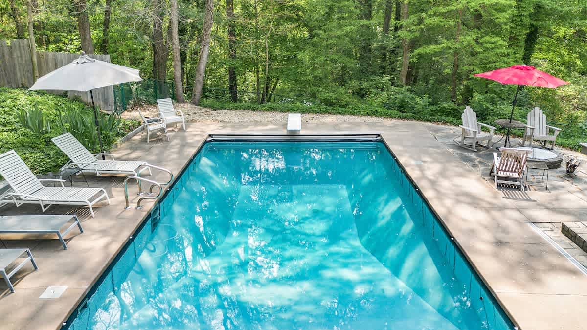 An inviting rectangular outdoor swimming pool with clear blue water, surrounded by a concrete deck furnished with lounge chairs, umbrellas, and Adirondack chairs. The area is bordered by a lush green forest.