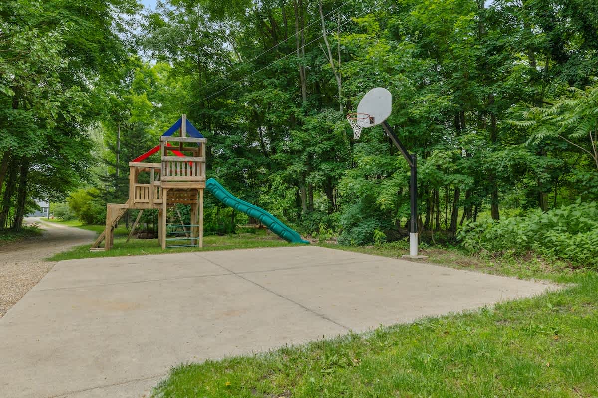A concrete pad serves as a half-court basketball area with a hoop, adjacent to a wooden playset featuring a slide. The area is surrounded by lush green grass and mature trees, suggesting a spacious backyard.