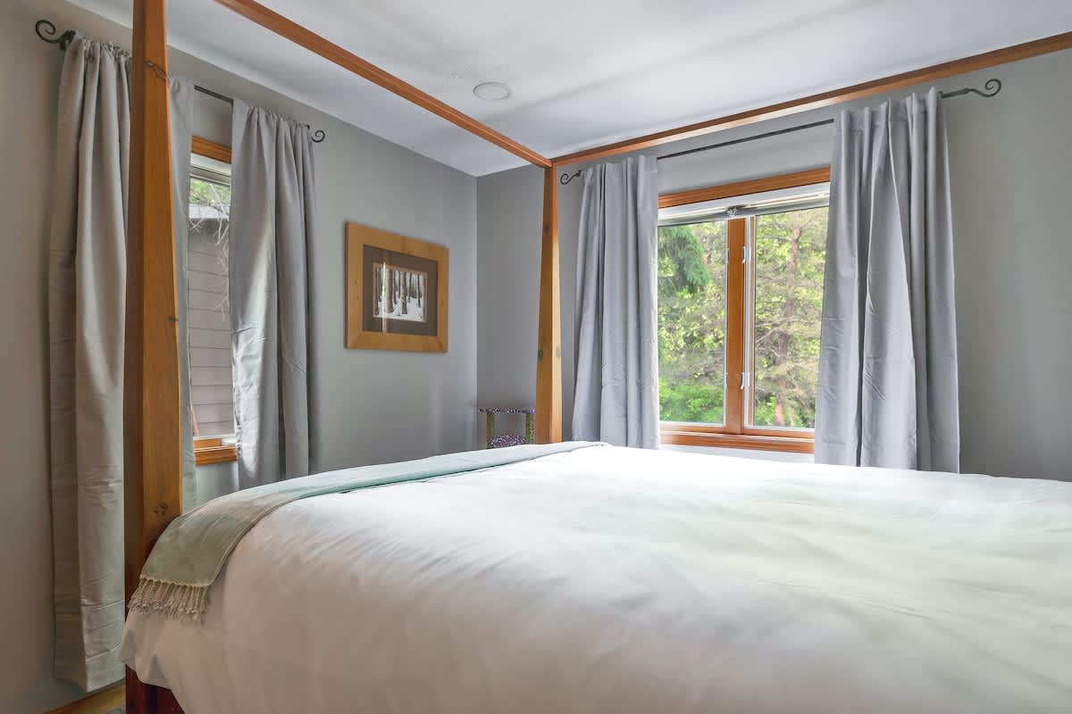 Bedroom with a wooden four-poster bed, two windows with light gray curtains, and a framed artwork on the wall, offering a bright and airy feel.