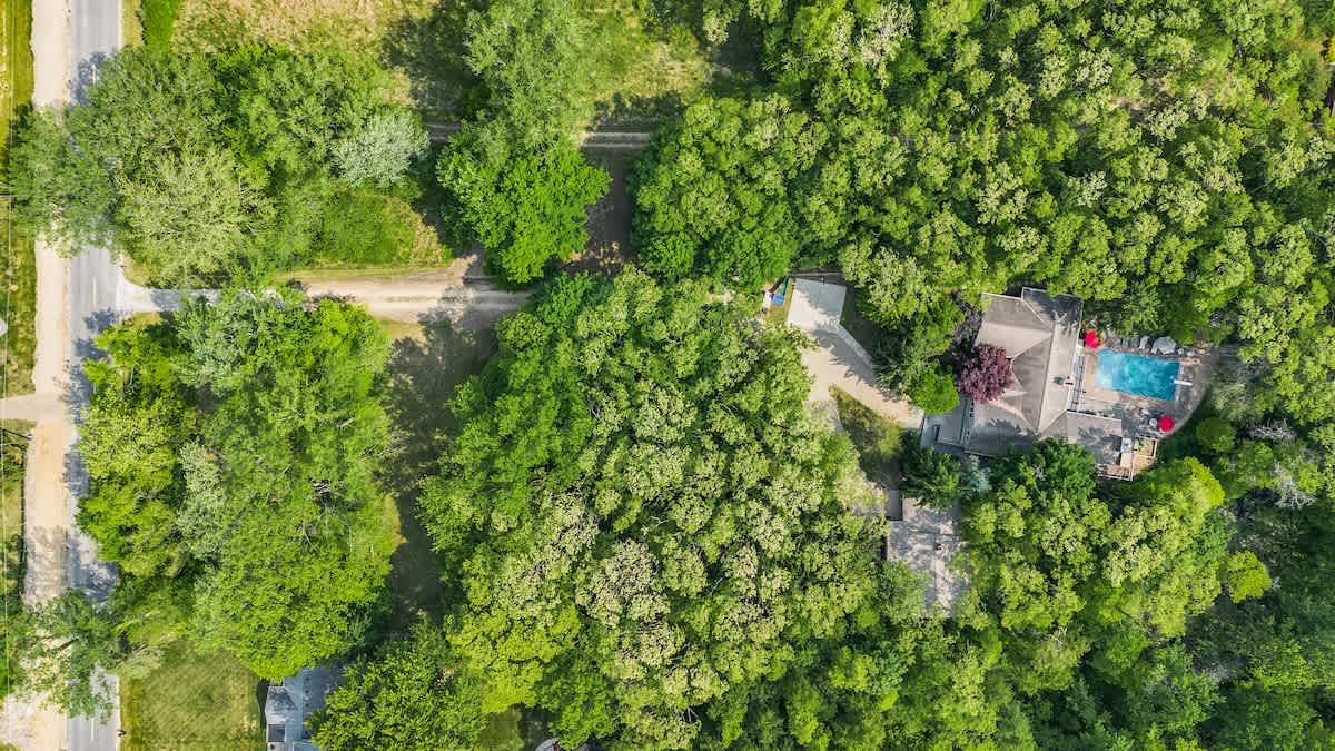 Aerial view of a house nestled in dense green trees, featuring a blue swimming pool with red umbrellas on a patio, and a winding driveway leading to the property. A road is visible on the left.