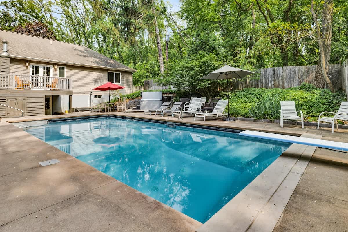 An inviting outdoor swimming pool with a diving board, surrounded by concrete decking, lounge chairs, and umbrellas, with a multi-level house and lush green trees in the background.