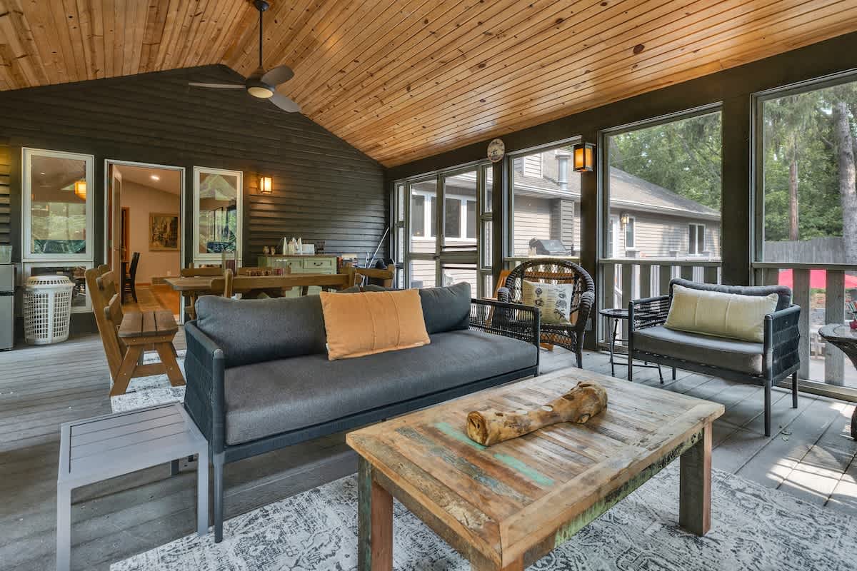 The lounge area of the screened porch features a dark gray sofa, a rustic wooden coffee table, and various wicker seating options, all set on a patterned outdoor rug. The vaulted wood-paneled ceiling and large windows create an airy feel.