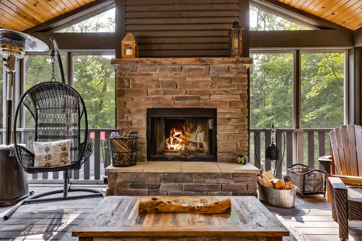 A detailed view of the stone fireplace in the screened porch, featuring a burning fire and rustic decor. A hanging egg chair and a patio heater are visible nearby, set against a backdrop of lush greenery outside the windows.