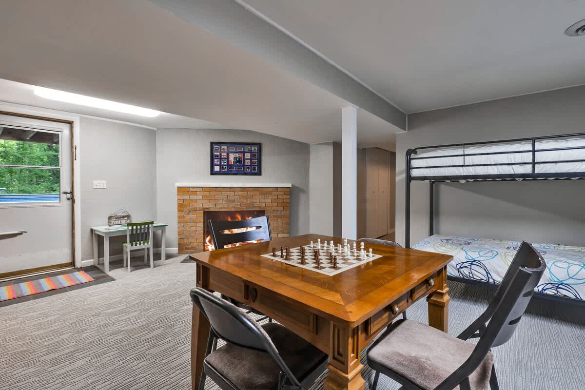 A view of the basement game room with a brick fireplace, a wooden game table with a chess set, and metal bunk beds. An exterior door with a small rug is visible, along with a small children's table and chairs.
