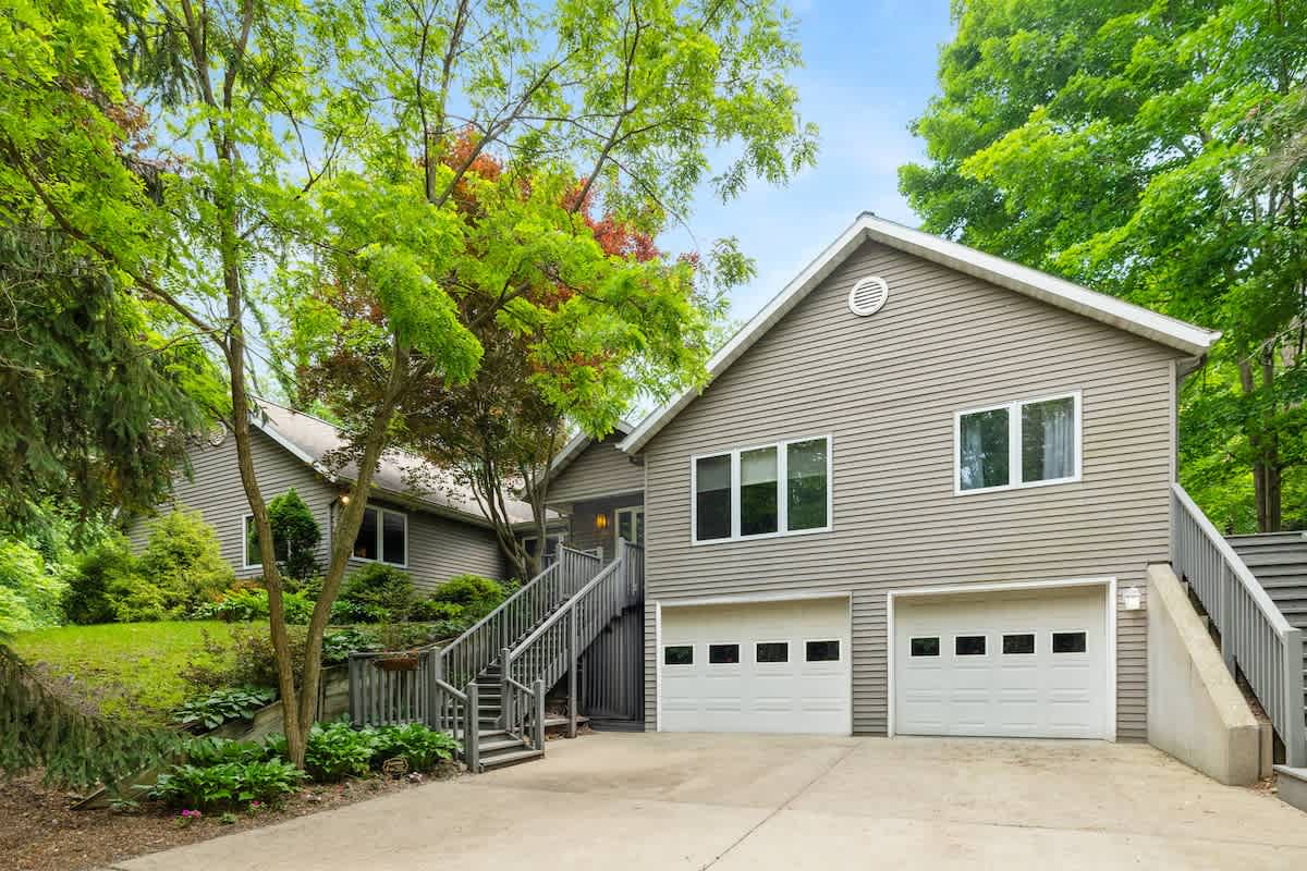 The front exterior of a multi-level house with light grey siding, featuring two garage doors, a concrete driveway, and multiple sets of stairs, all surrounded by mature green trees.